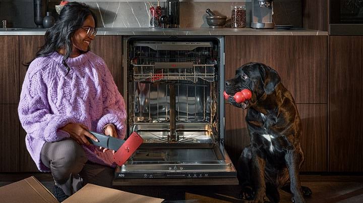 Kvinne pakker opp reservedeler for oppvaskmaskinen mens hun ler mot en svart hund som har en leke i munnen