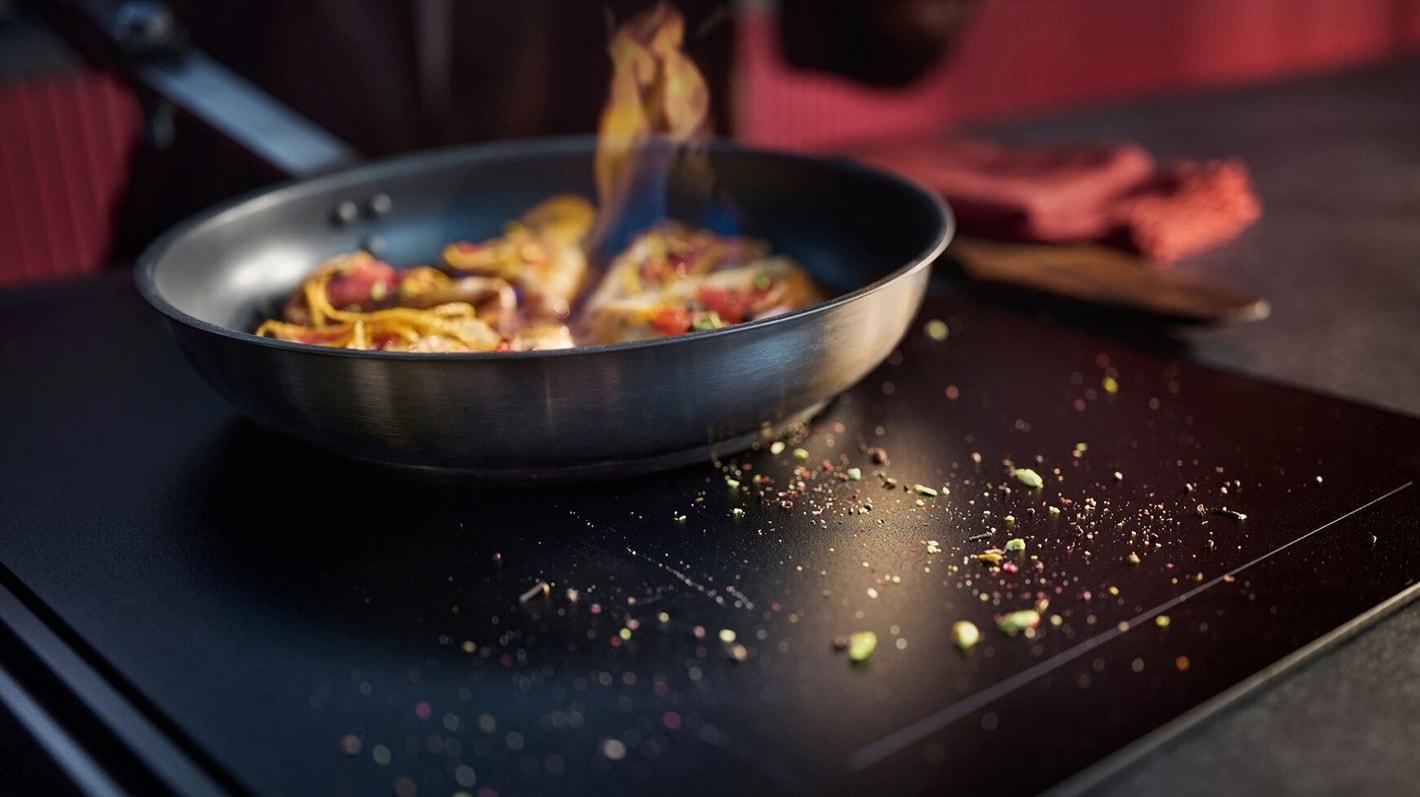 En person lager mat i en stekepanne på en platetopp. Rundt ligger det diverse smuss og krydderrester.