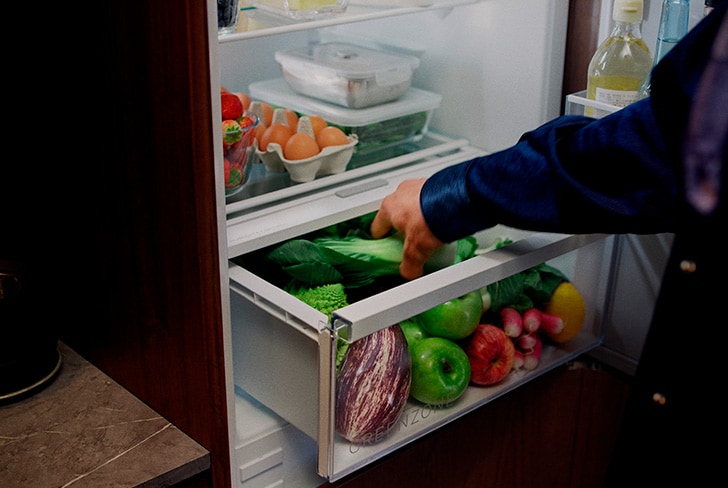 Åpent kjøleskap med frukt og grønnsaker i grønnsaksskuffen og en person som organiserer innholdet.